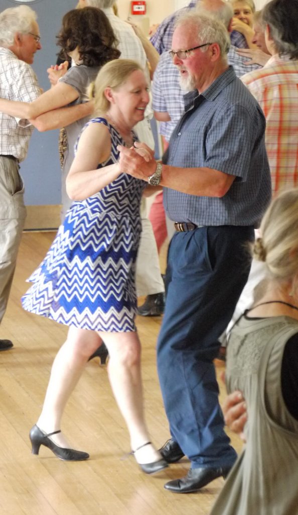 Couple in the foreground dancing in waltz hold, several other dancers visible in the background.