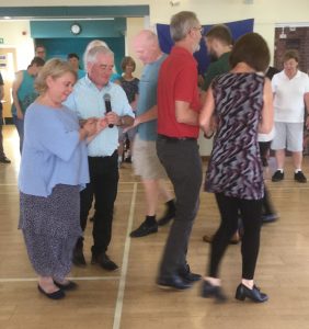 Four couples dancing around in cross hand hold, other people watching in the background.